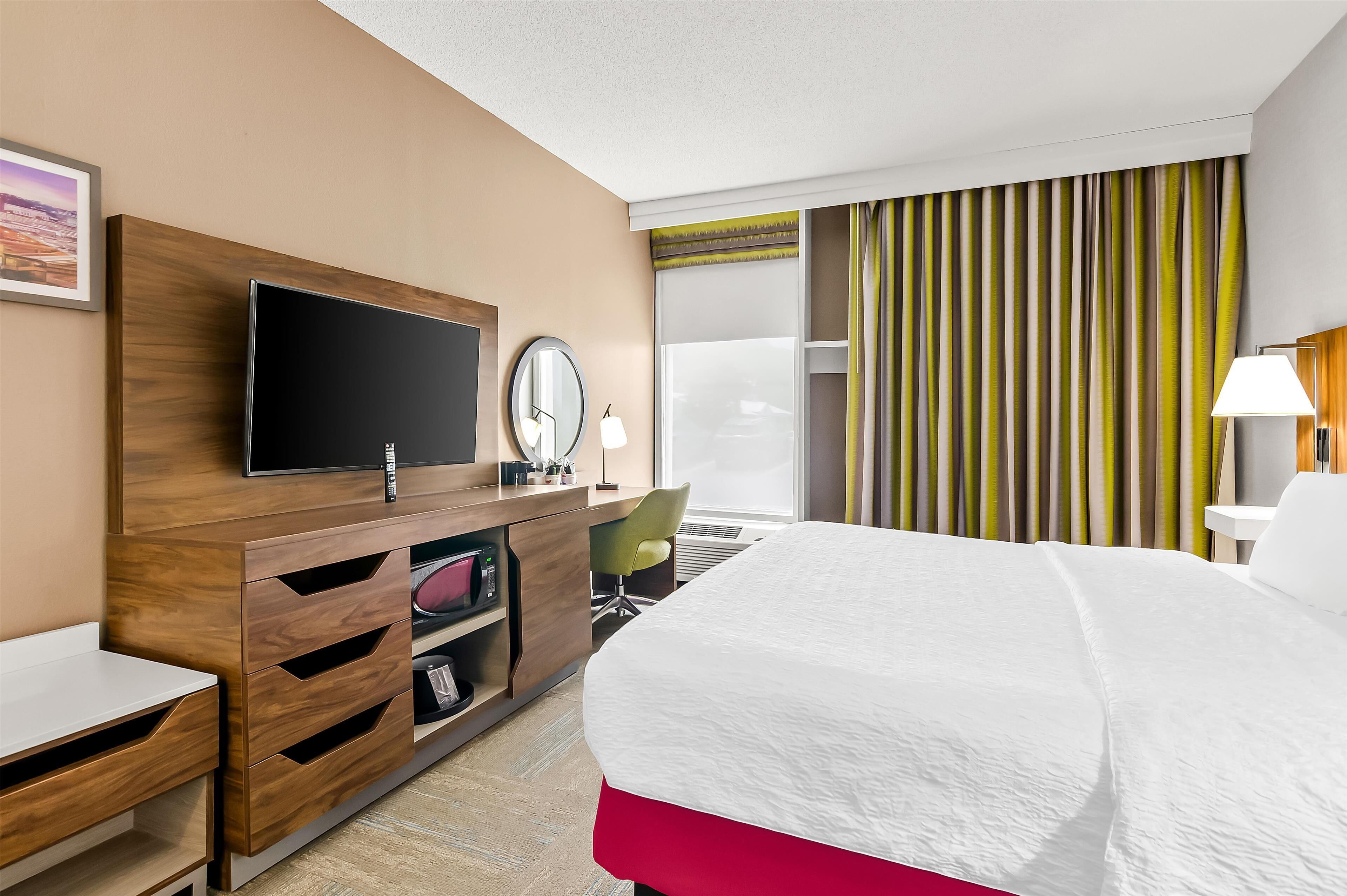 A hotel room with a bed, a desk with a TV, a chair, and a window with curtains.