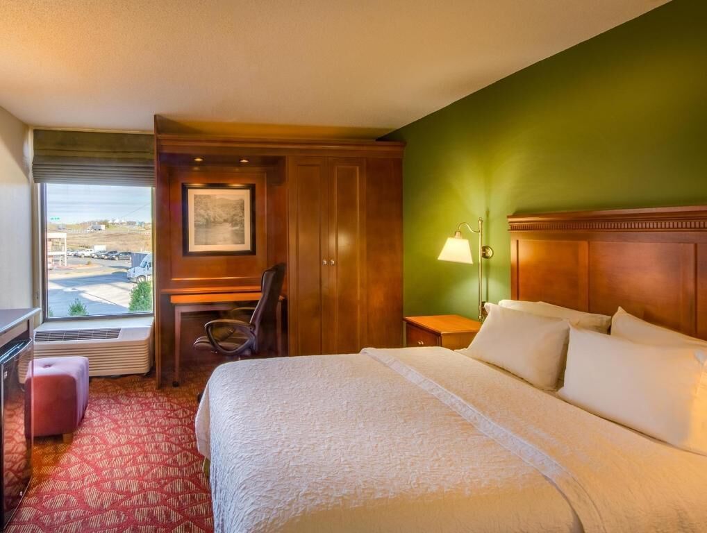A hotel room with a bed, a desk, a chair, a lamp, and a window with a view.
