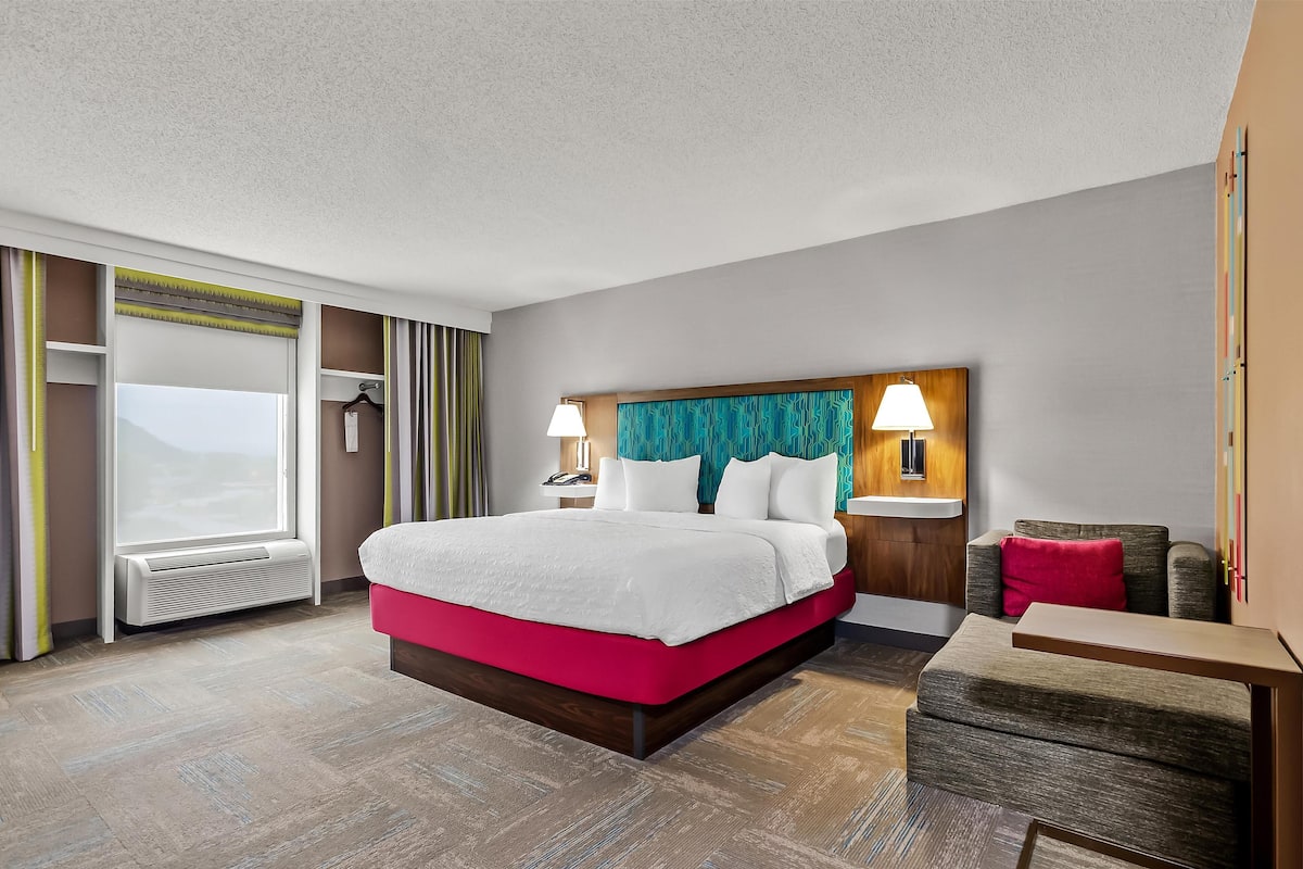 Ein Hotelzimmer mit einem großen Bett, einem Schreibtisch, einem Sessel, einem Nachttisch, einer Lampe und Blick ins Freie.