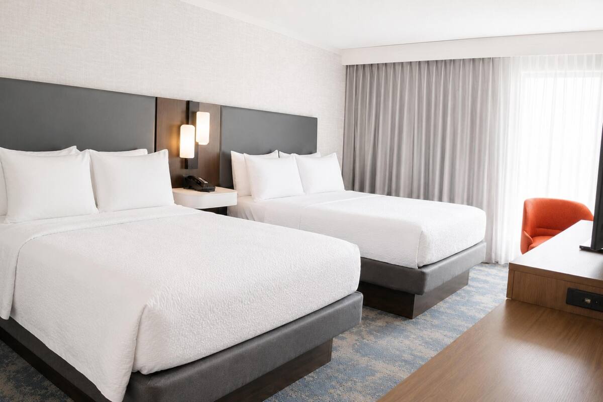 A hotel room with two beds, a desk, a chair, a nightstand, and a window with curtains.