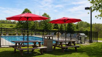 A fenced outdoor area with picnic tables and red umbrellas, a pool, and a barbecue grill.