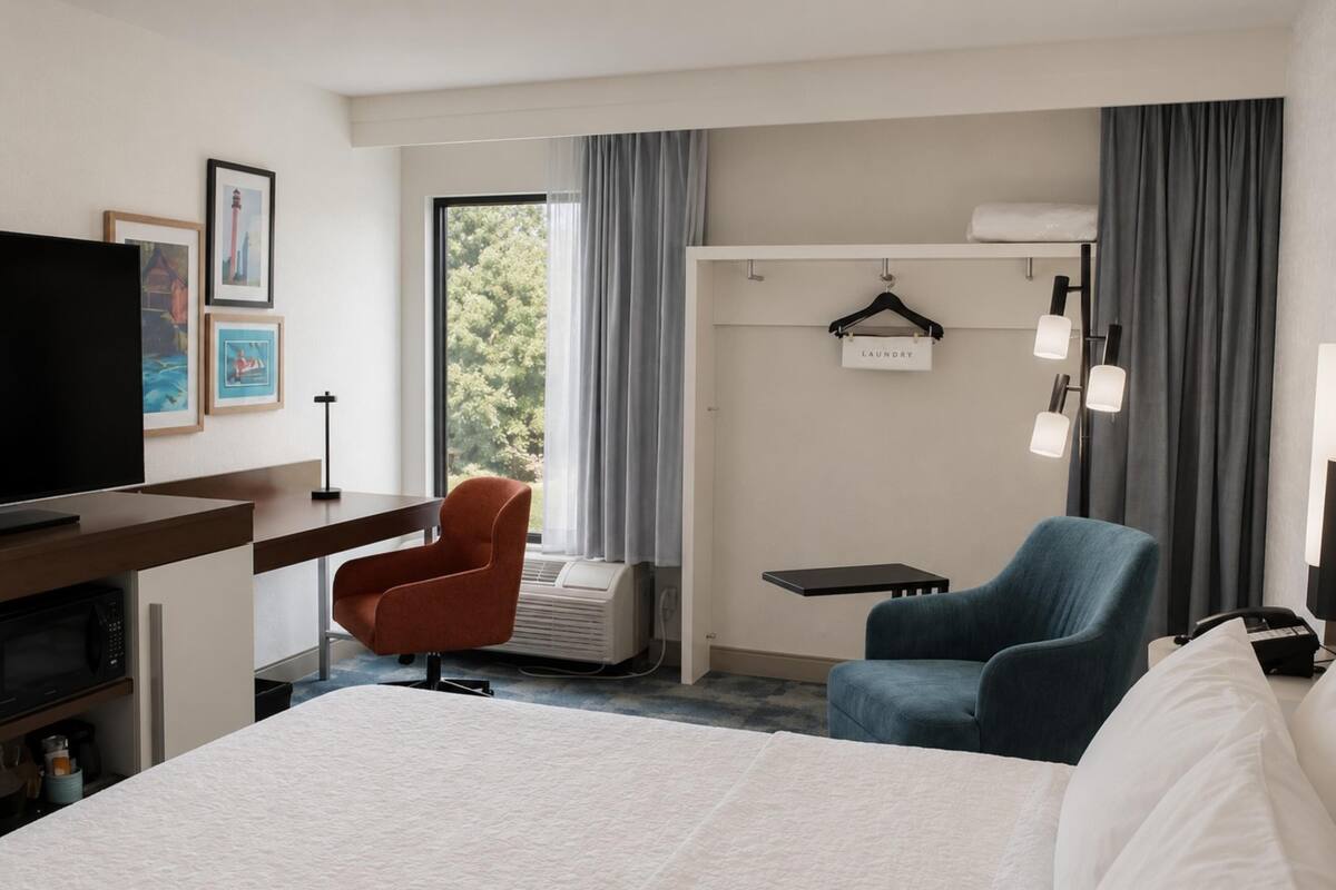 A hotel room with a bed, a desk, a television, a chair, a hanging laundry sign, and a window with curtains.