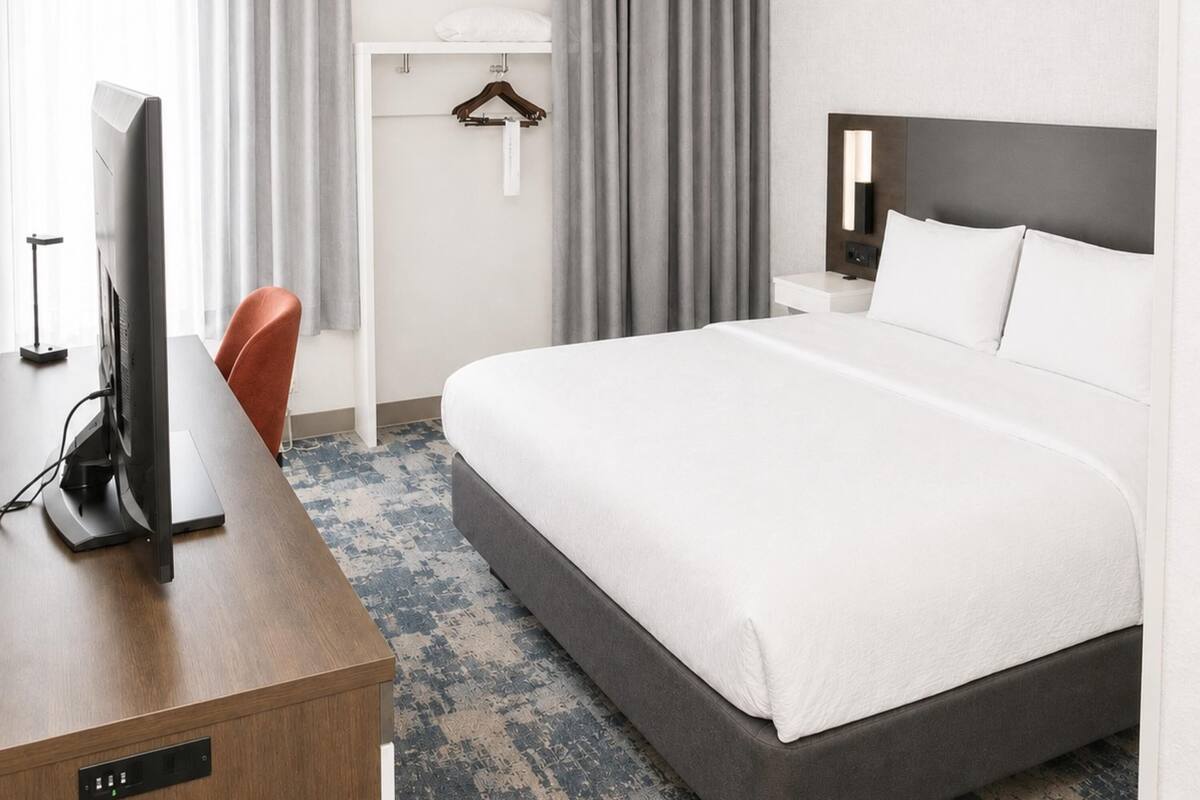 A hotel room with a bed, a desk with a computer, a chair, a bedside table, and a hanging light fixture.