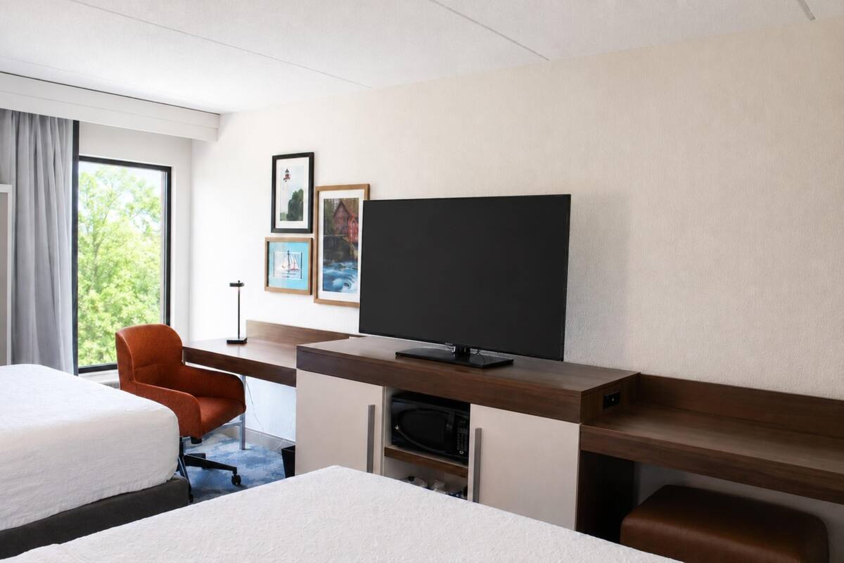 A hotel room with a bed, a desk with a television, a chair, and a window with curtains.