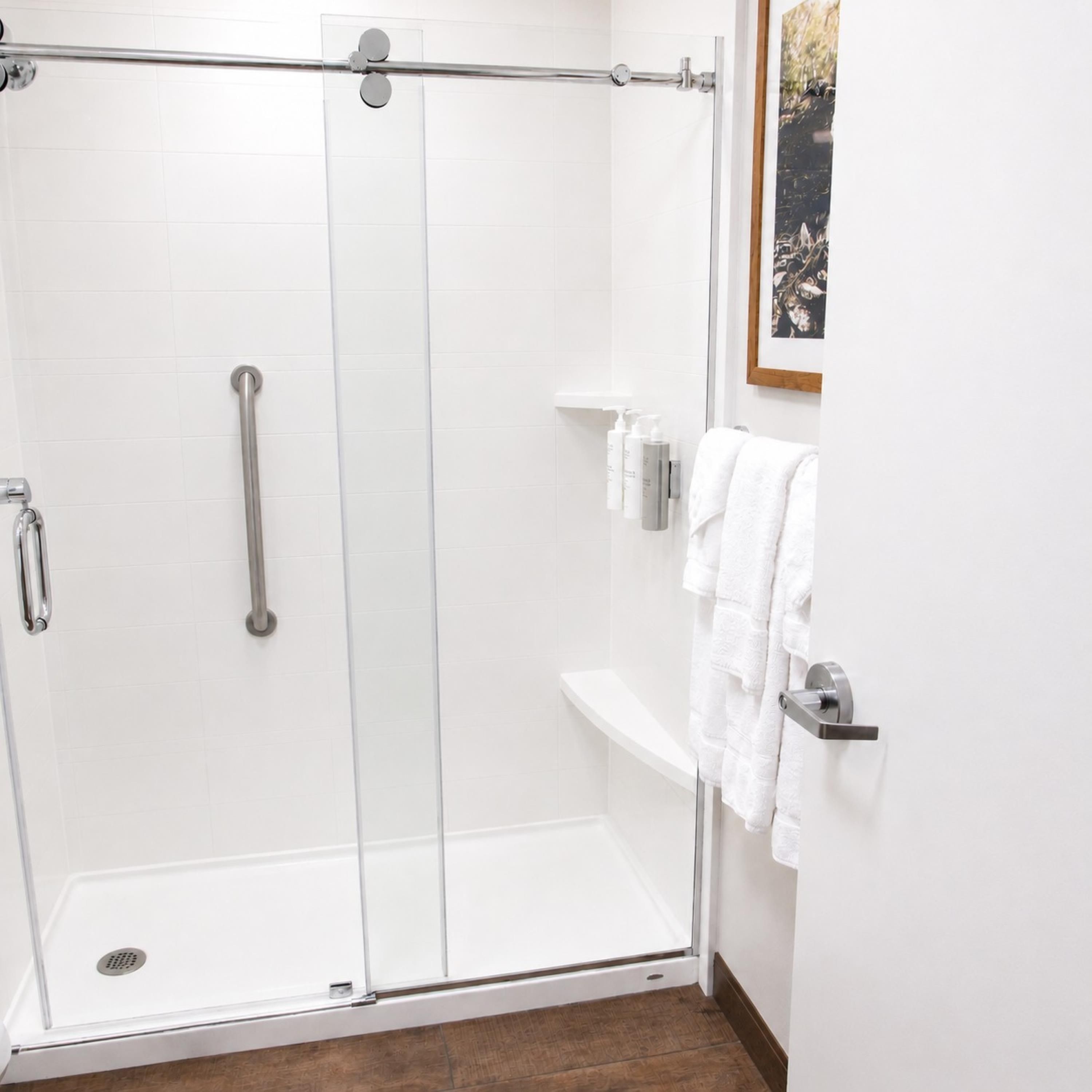 A glass-enclosed shower with a handrail, a white tiled wall, a wooden floor, and a framed picture.