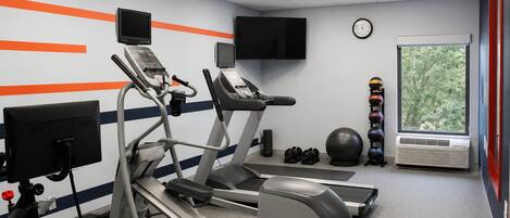 A gym with treadmills, a stationary bike, a rowing machine, a wall-mounted TV, a clock, a window with a view of trees, and a set of dumbbells.