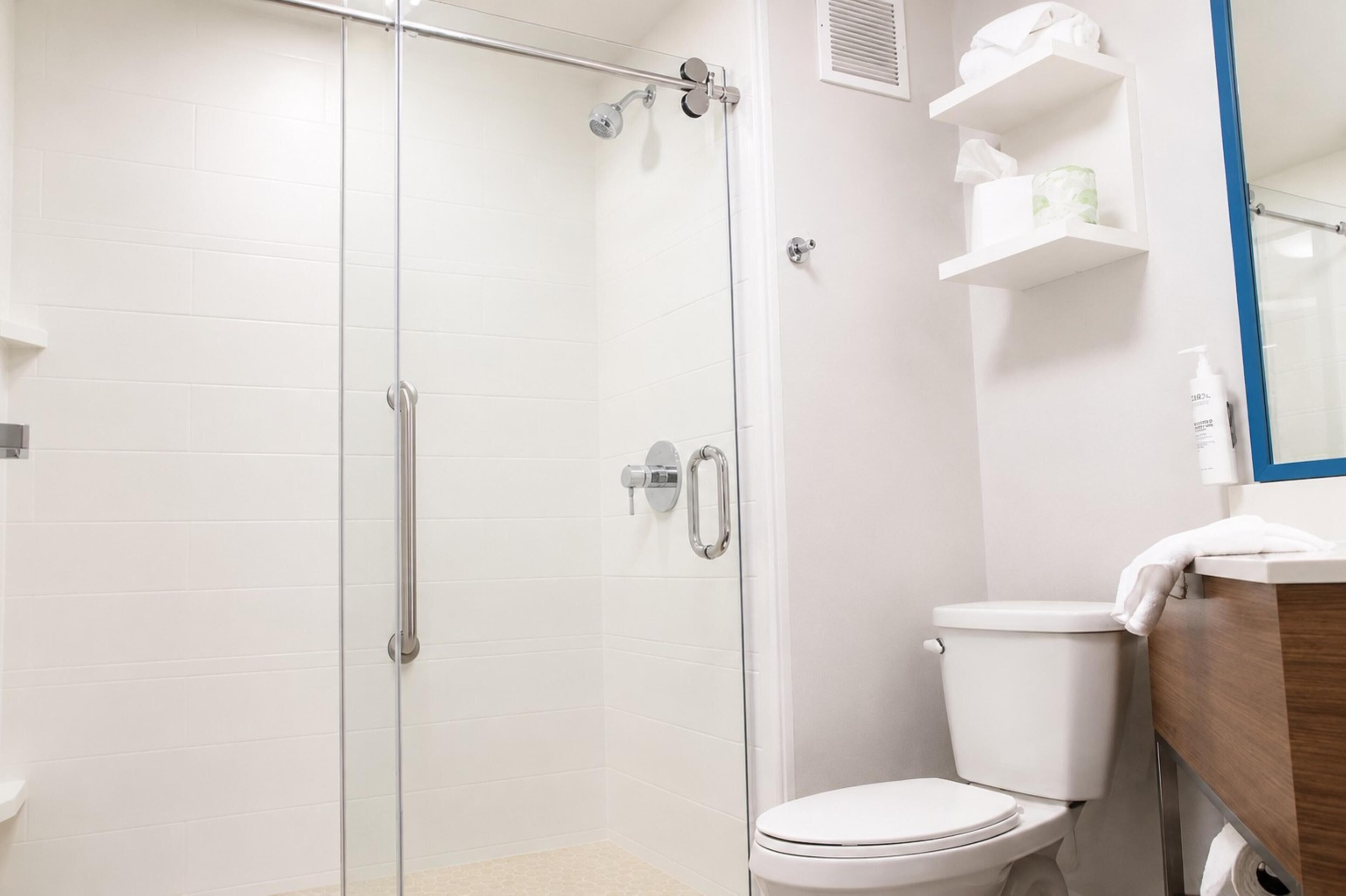 Un baño con ducha envuelta en vidrio, inodoro blanco y tocador de madera con espejo.