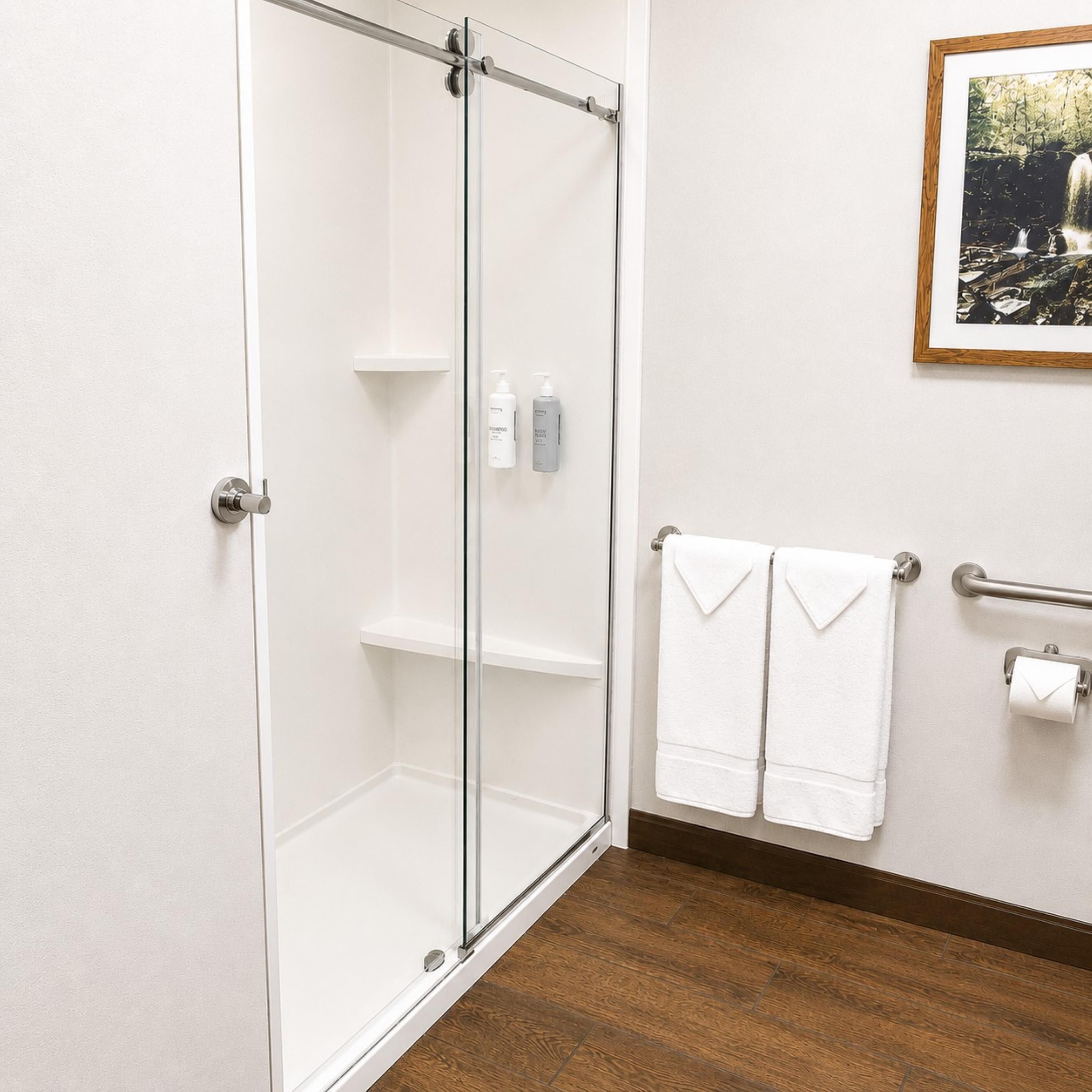 Een douche met een glazen deur, een houten vloer, een handdoekhouder met twee handdoeken en een ingelijst schilderij aan de muur.