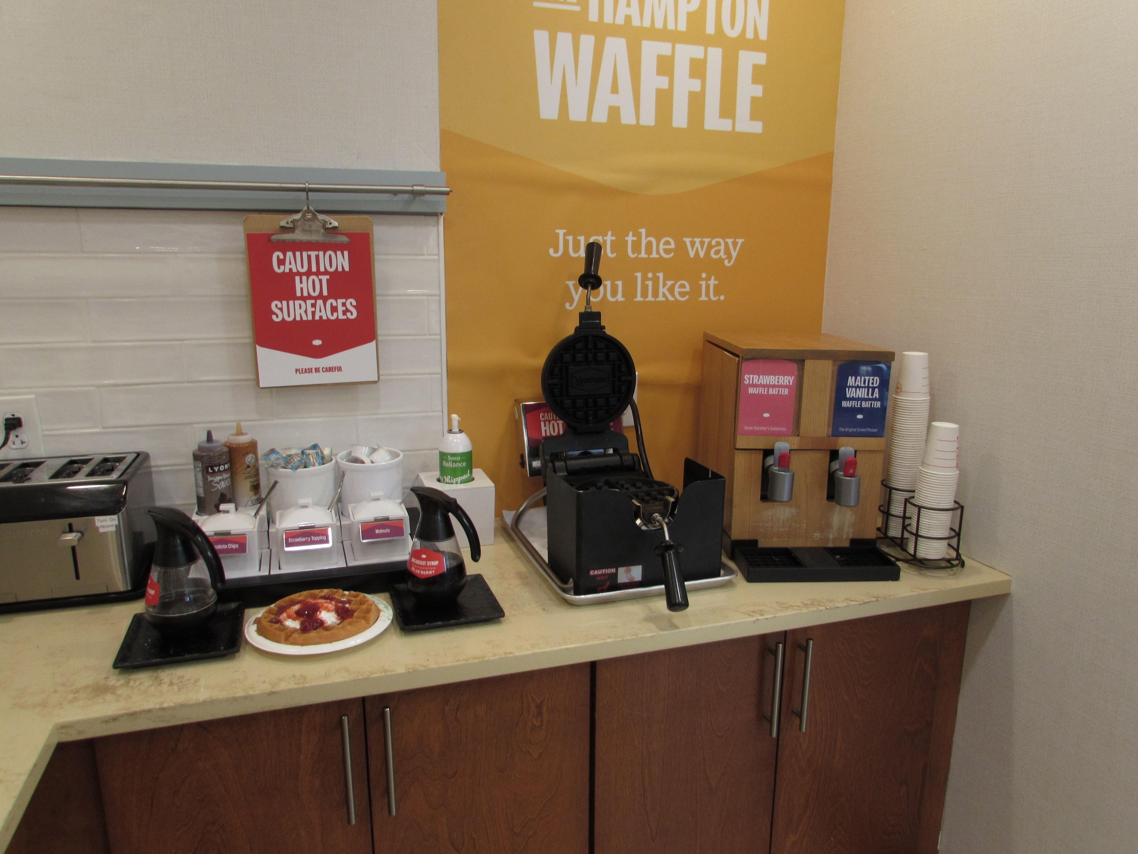 A kitchen counter with a waffle maker, waffle batter dispensers, and a caution sign for hot surfaces.