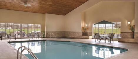 An indoor swimming pool with a wooden ceiling, clear glass windows, and outdoor seating area.