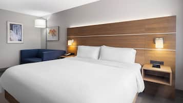 Quarto de hotel moderno com uma cama grande, uma mesinha de cabeceira com abajur, uma cadeira azul e obras de arte nas paredes.