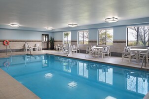 Una piscina interior con agua azul cristalina, rodeada de sillas y mesas blancas, y grandes ventanales que ofrecen vistas al exterior.