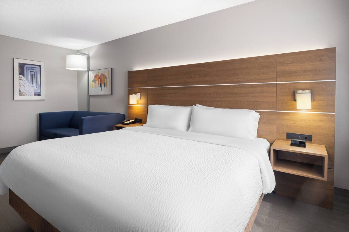 A modern hotel room with a large bed, a bedside table with a lamp, a blue chair, and artwork on the walls.