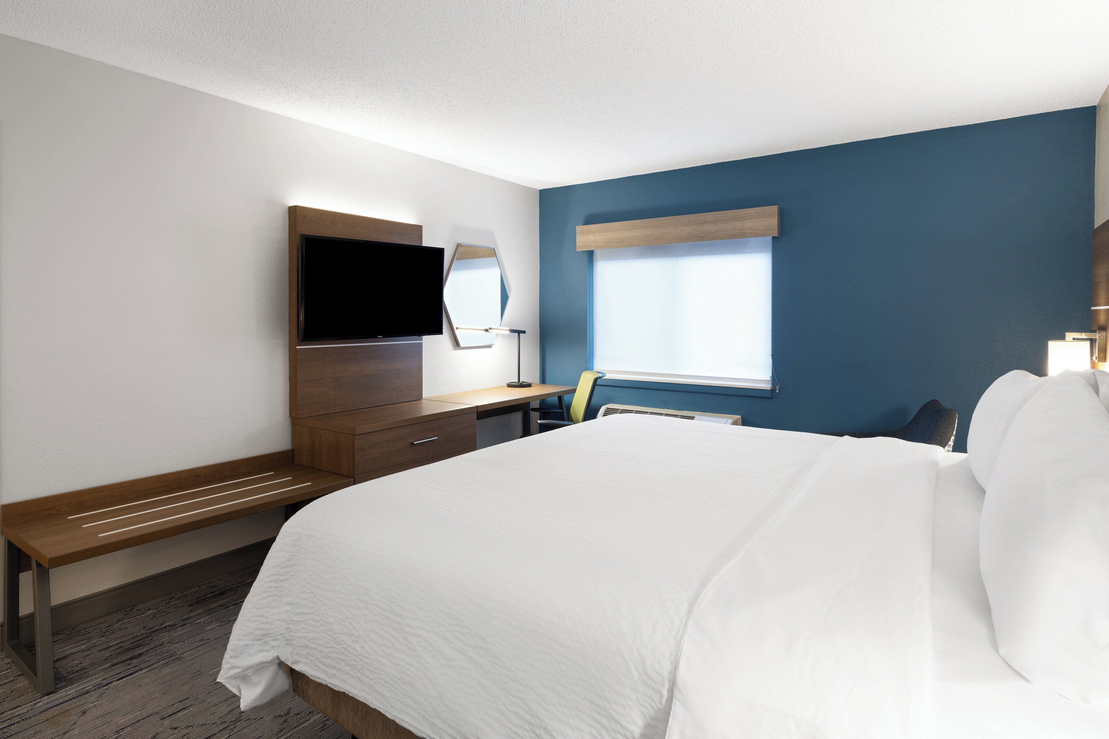 A hotel room with a large bed, a desk with a chair, a flat-screen TV, a mirror, and a window with blinds.