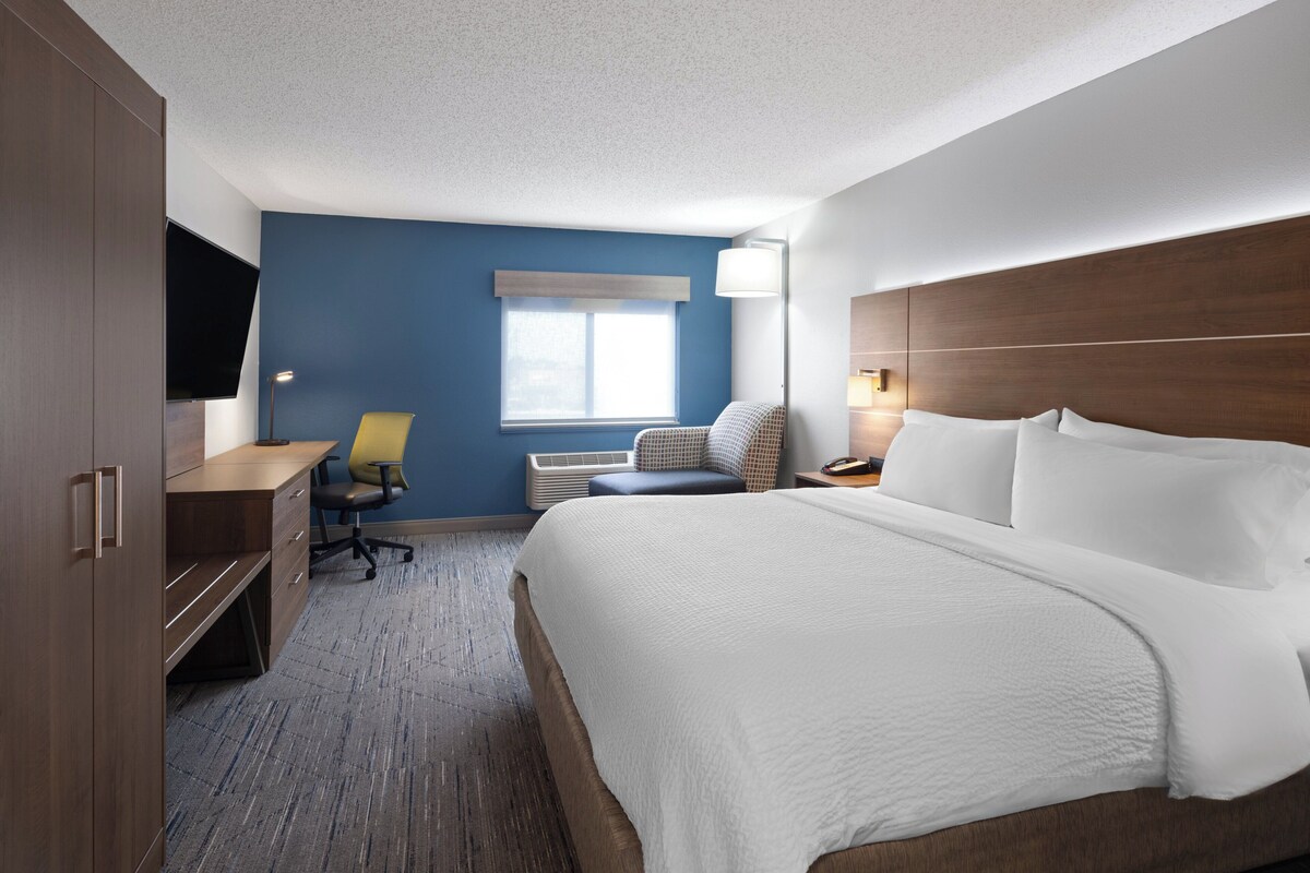 A hotel room with a large bed, a desk, a chair, a lamp, and a window.