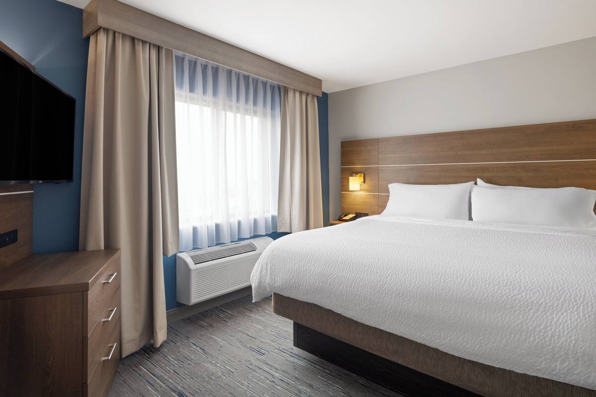 Ett hotellrum med en stor säng, en platt-TV, ett skrivbord med lådor, ett fönster med gardiner och en väggmonterad lampa.