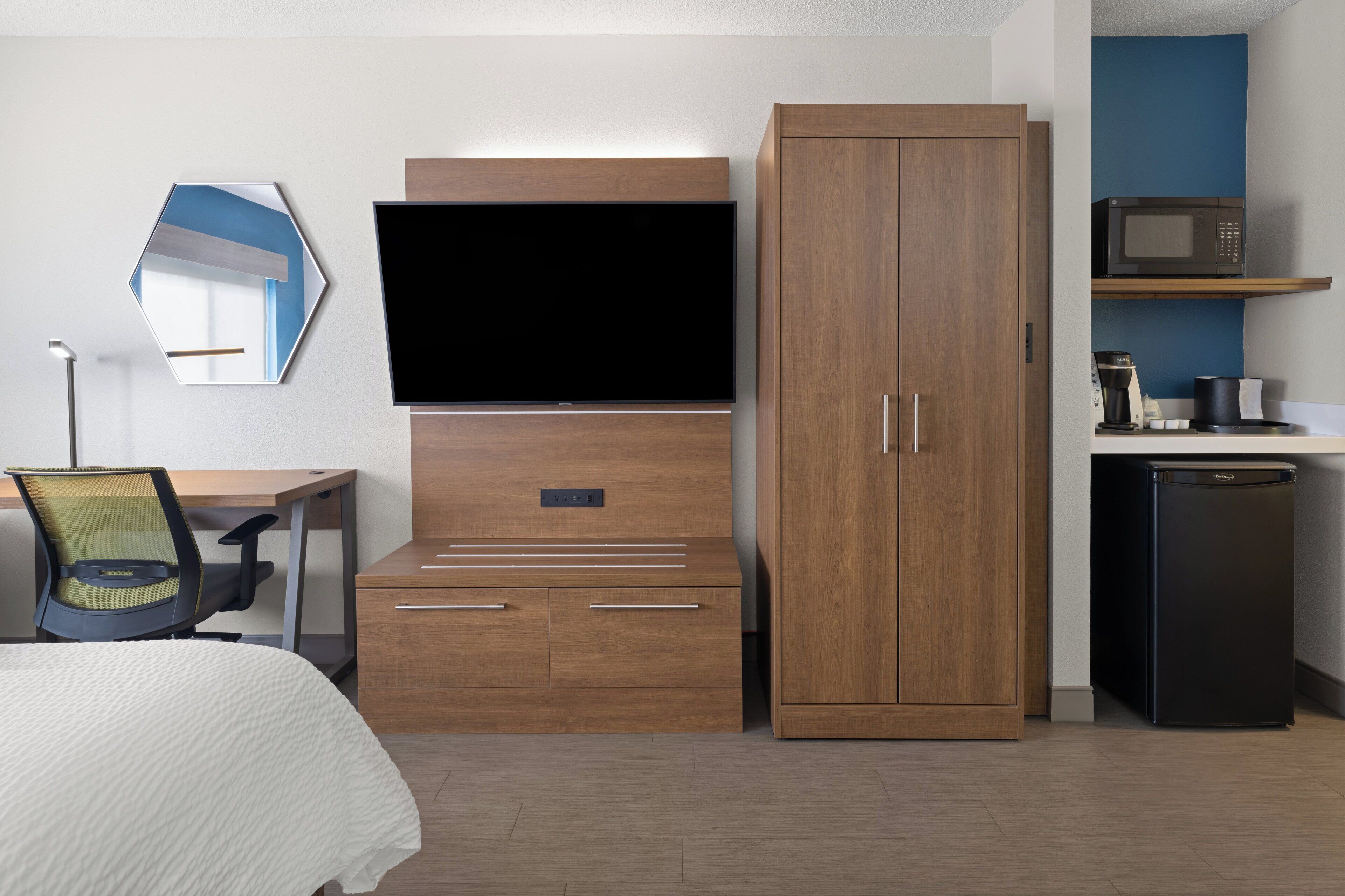 Quarto de hotel com cama, escrivaninha com cadeira, televisão, guarda-roupa, micro-ondas e frigorífico.