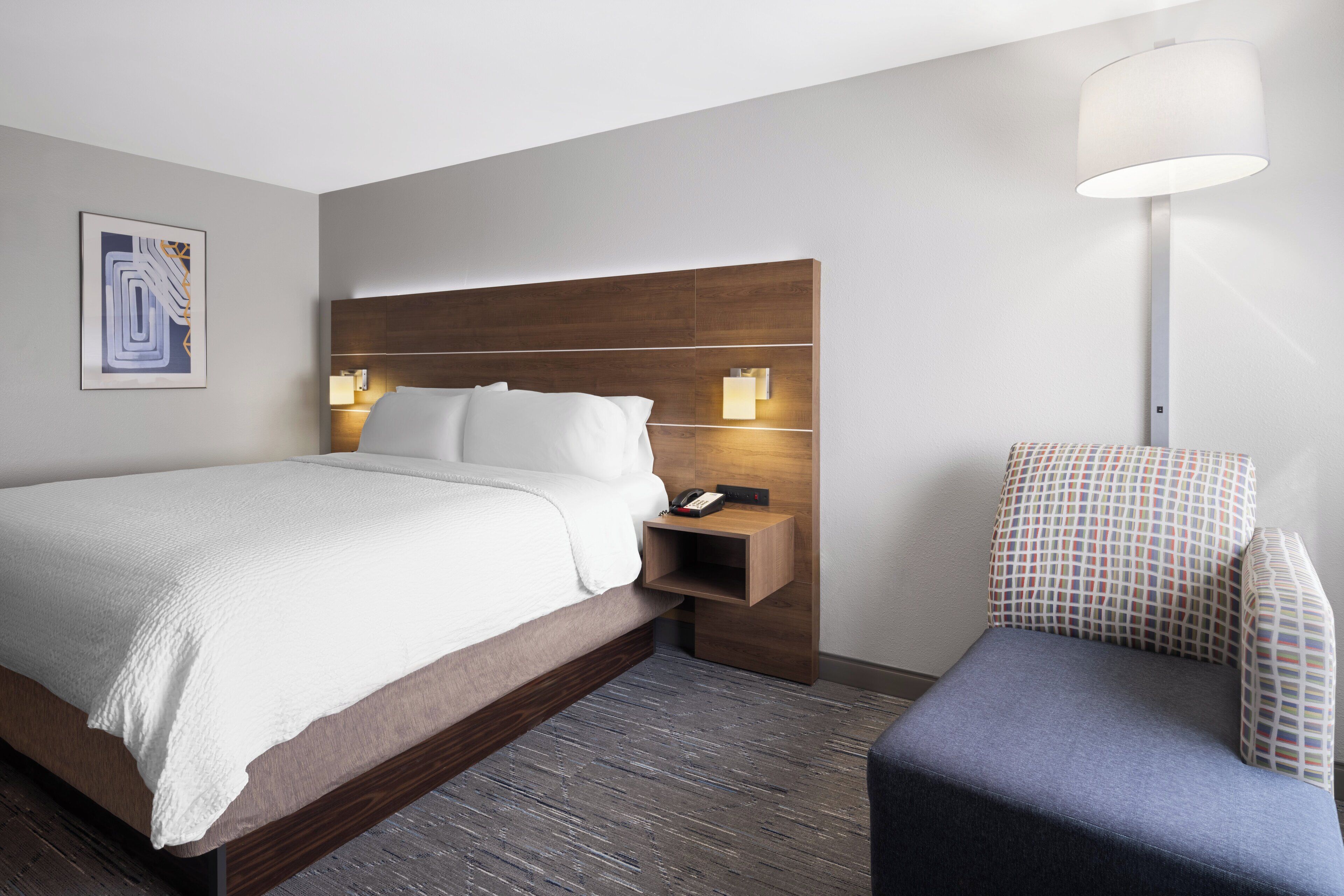 Una habitación de hotel moderna con cabecera de madera, una cama grande con sábanas blancas, una mesita de noche con lámpara y un sillón con estampado.