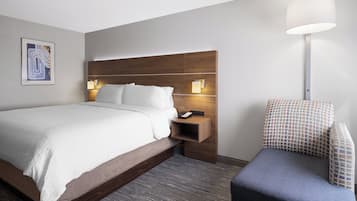Una habitación de hotel moderna con cabecera de madera, una cama grande con sábanas blancas, una mesita de noche con lámpara y un sillón con estampado.