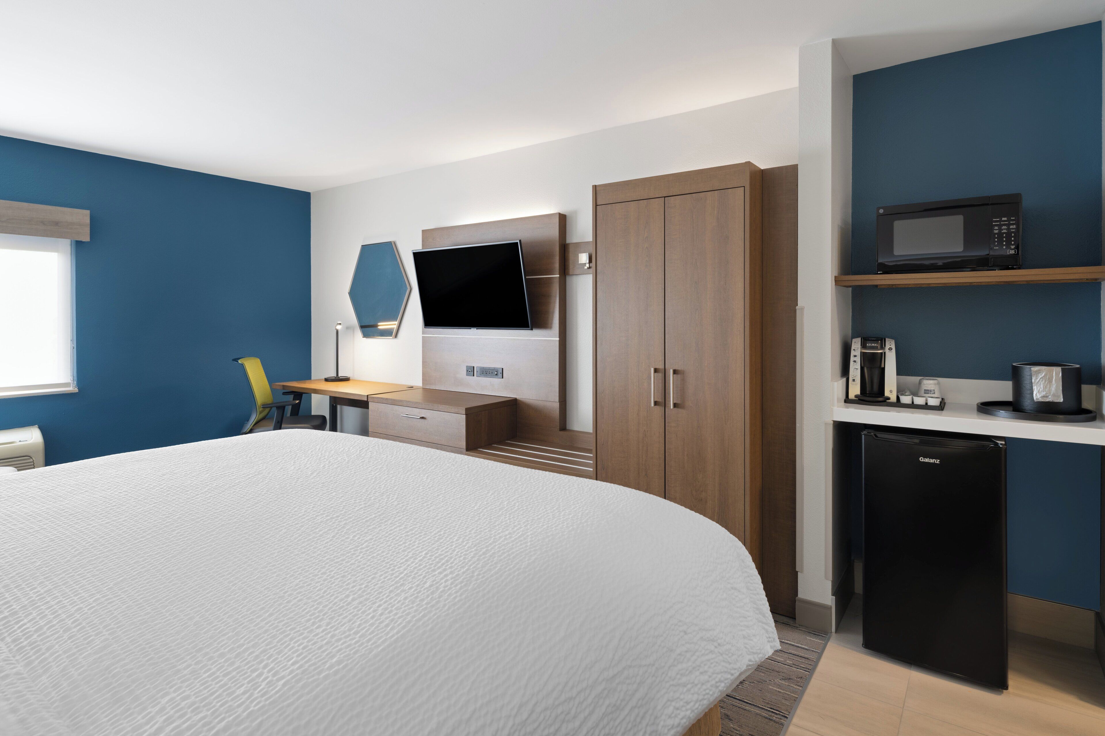 A modern hotel room with a large bed, a desk with a chair, a TV, a wardrobe, a mini-fridge, and a microwave on a shelf.