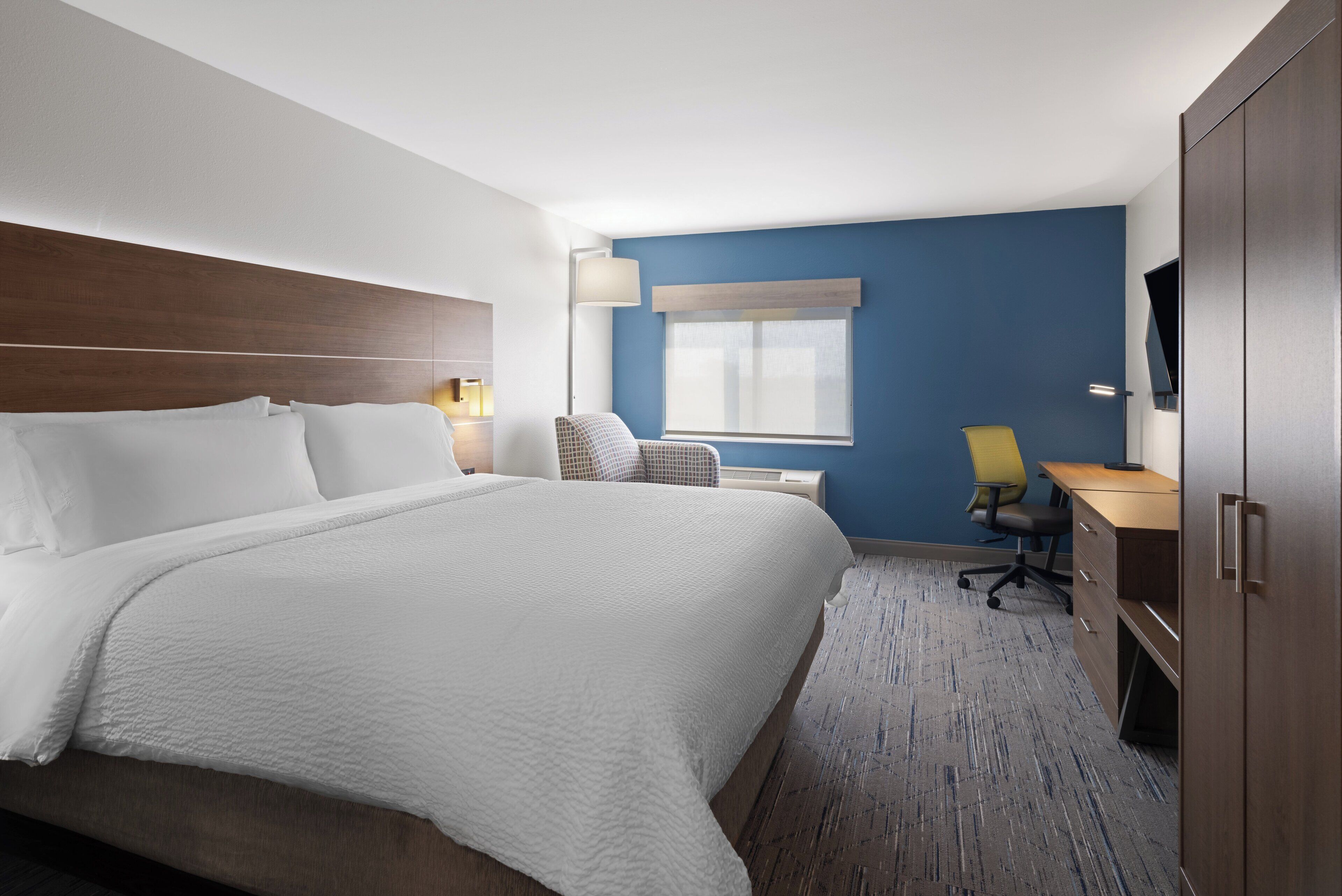 A hotel room with a large bed, a desk, a chair, a TV, and a window with blinds.