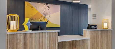 A hotel reception desk with a modern design, featuring a wooden panel backdrop, a large abstract painting, and two illuminated wall sconces.