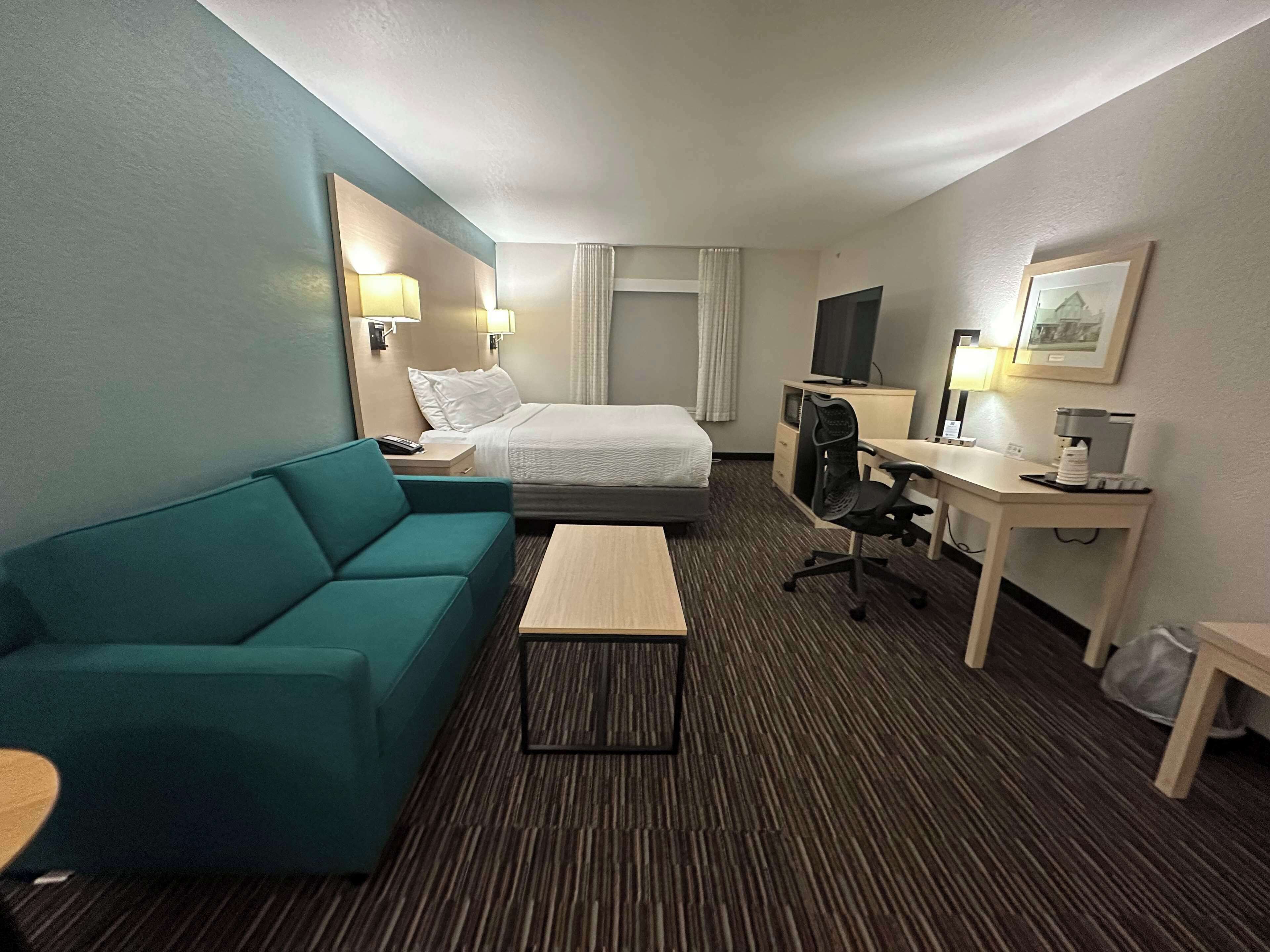 A hotel room with a bed, a teal sofa, a desk with a computer, a coffee maker, and a TV.