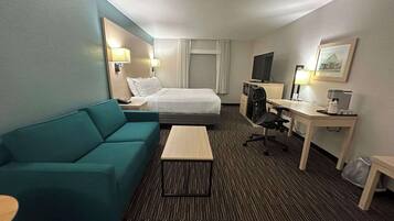 A hotel room with a bed, a teal sofa, a desk with a computer, a coffee maker, and a TV.