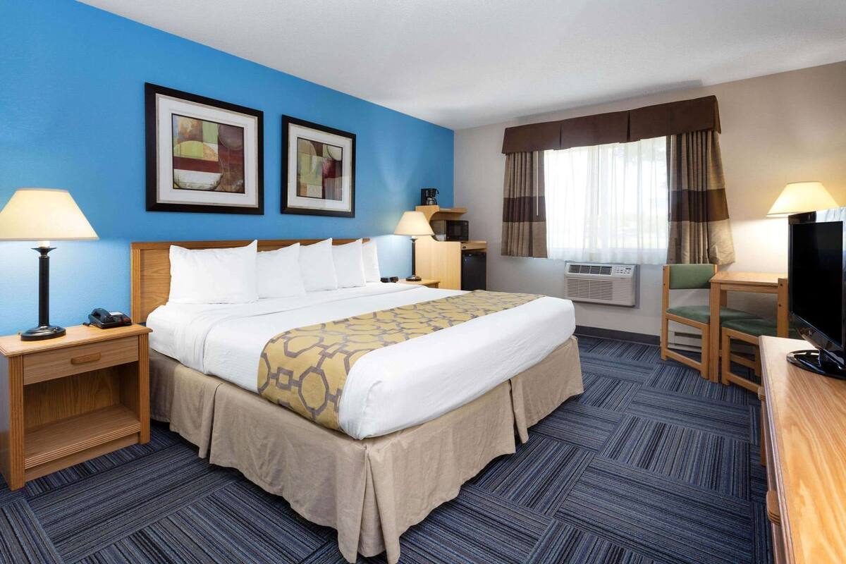 Een hotelkamer met een bed, nachtkastjes, een bureau, een stoel, een televisie, een raam met gordijnen en twee ingelijste foto's aan de muur.