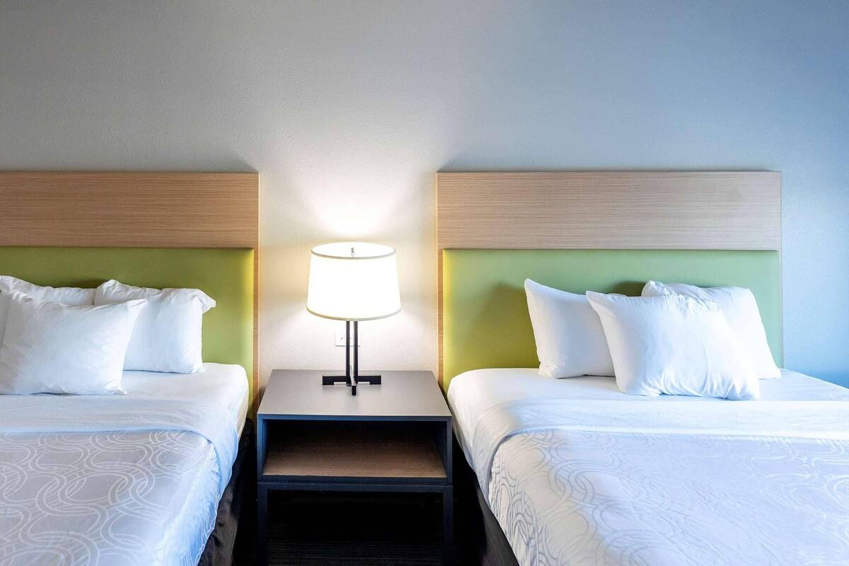 A hotel room with two single beds, each with white bedding and pillows, a nightstand with a lamp, and a green headboard.