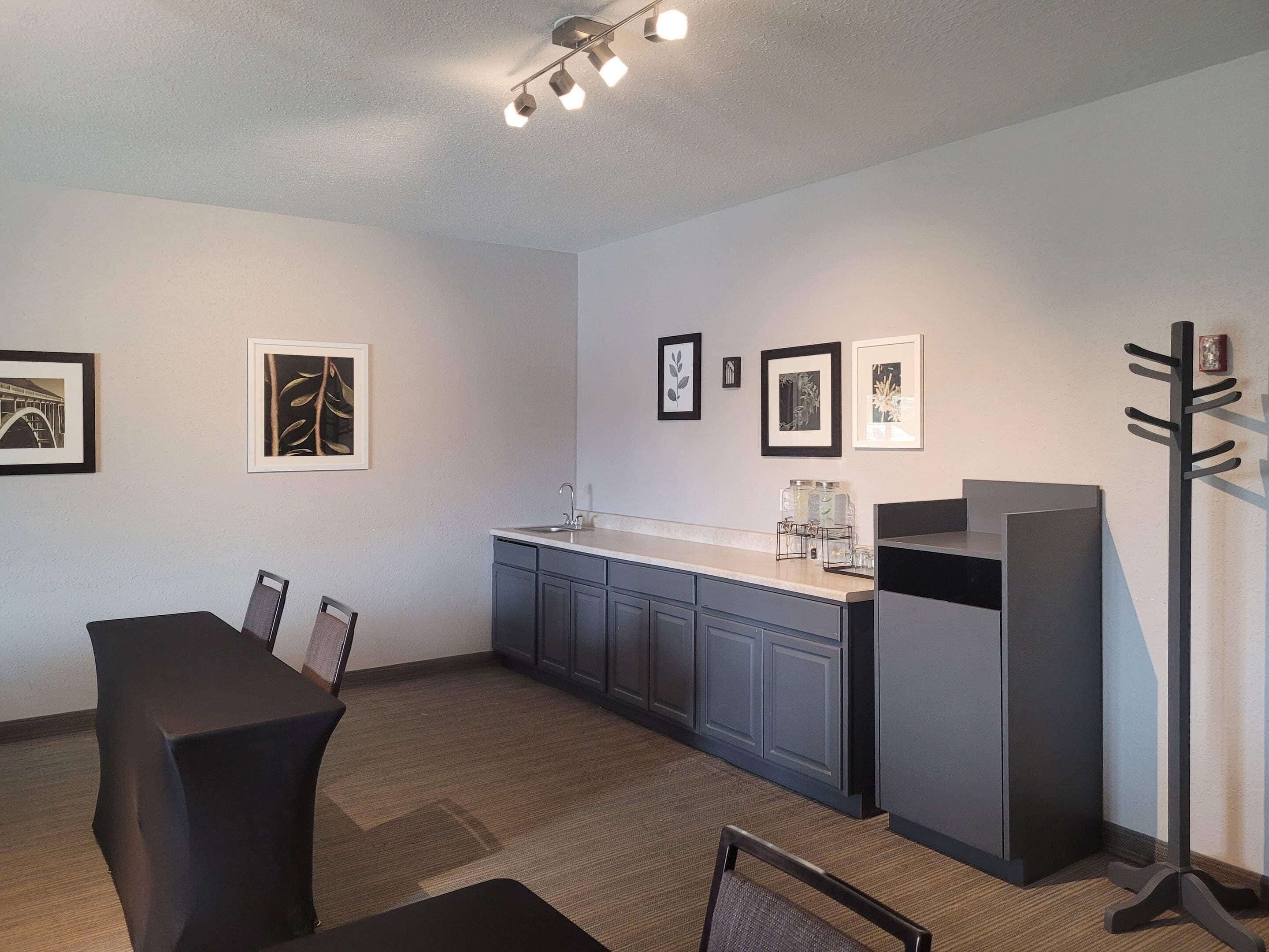 A modern kitchen with grey cabinets, a white countertop, and a black chair. There are framed pictures on the wall and a coat rack.