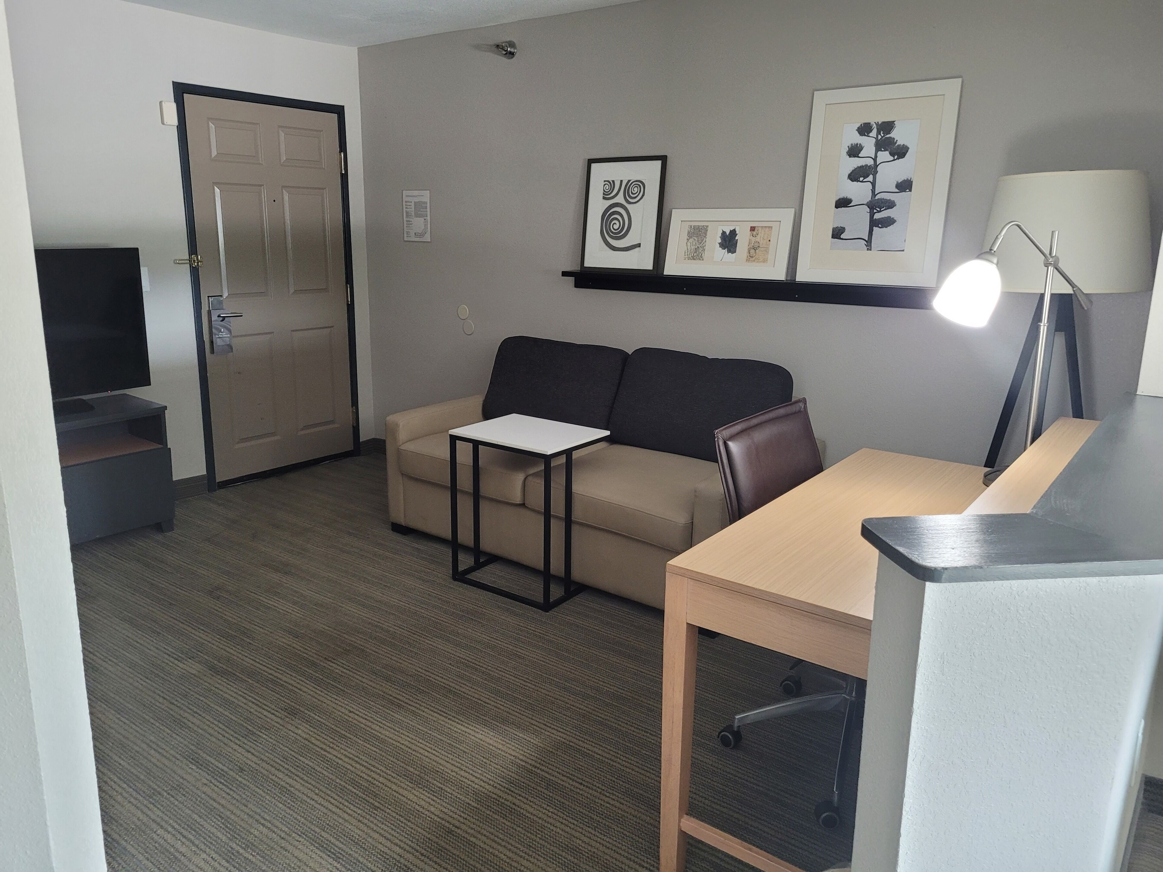 A compact living room with a sofa, a small table, a TV, and a desk with a lamp.