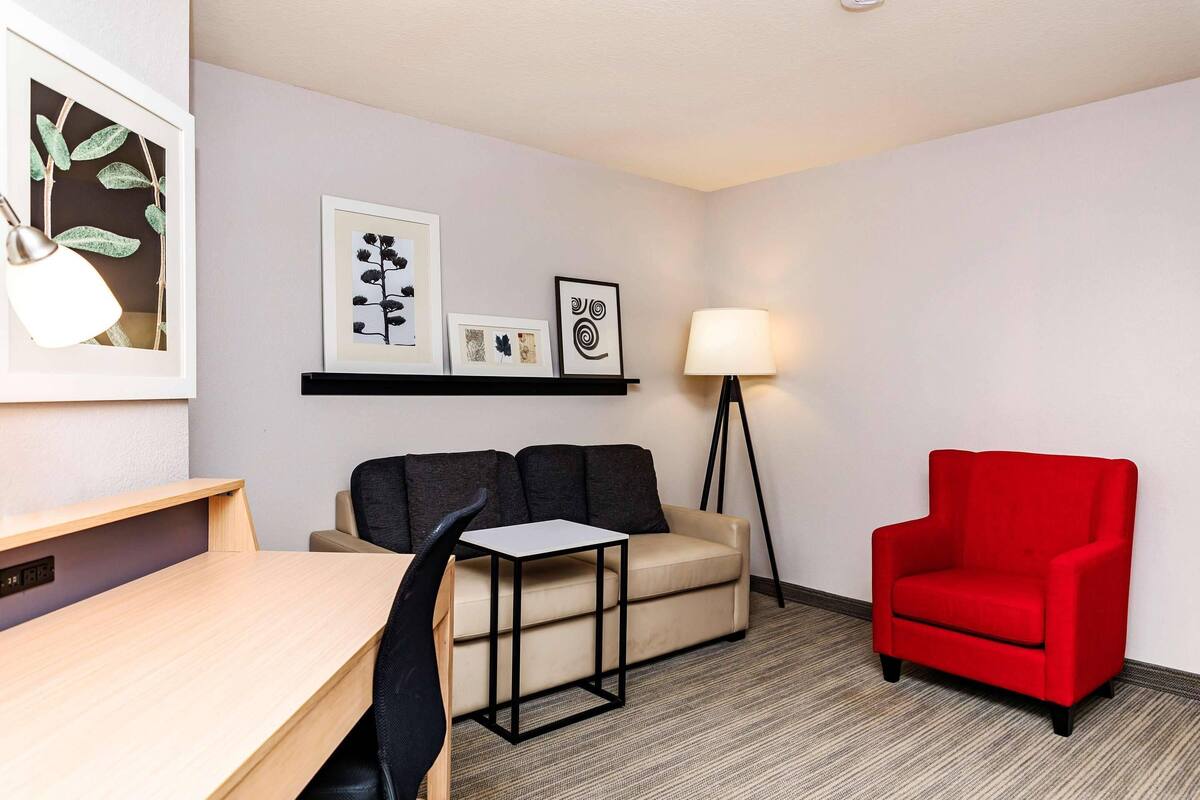 A hotel room with a desk, a sofa, a red armchair, a floor lamp, and wall art.