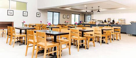A spacious dining area with multiple tables and chairs, a kitchen in the background, and framed artwork on the walls.