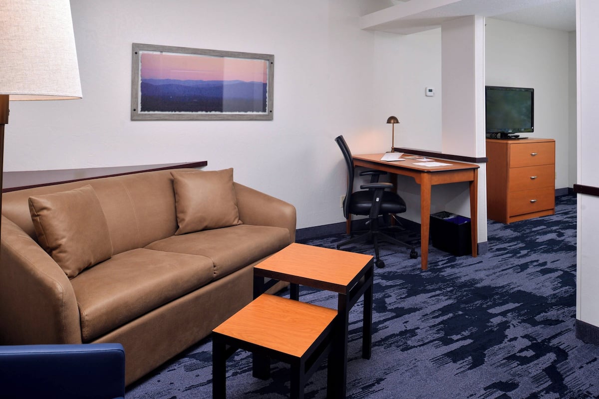 A hotel room with a brown leather sofa, a wooden desk with a chair, a small wooden table, a television on a cabinet, and a framed picture on the wall.
