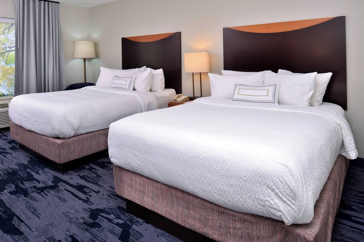 A hotel room with two beds, a large window with curtains, a lamp, and a wall-mounted artwork.