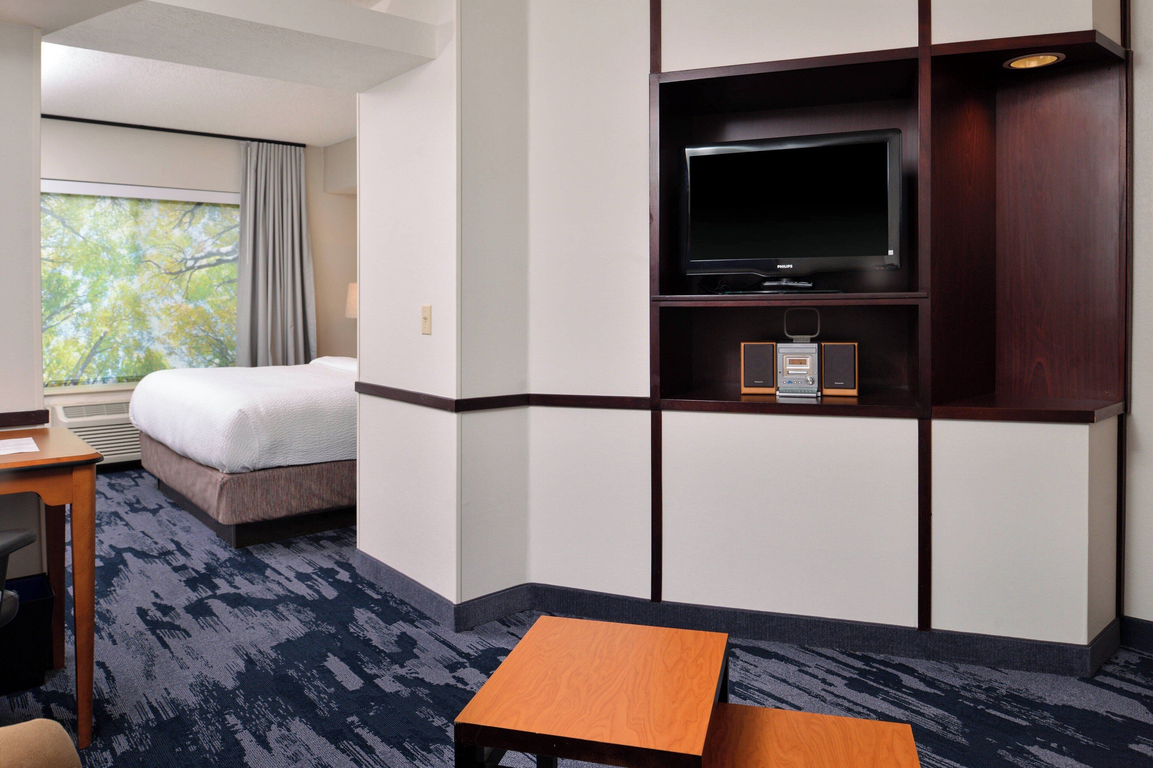 Habitación de hotel con cama, escritorio, televisor y ventana con vistas a árboles.