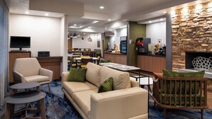 Un moderno vestíbulo de hotel con un sofá color beige, una silla de madera, una mesita y una cuna para bebé. Se ve un televisor, una zona de cocina y una chimenea de piedra al fondo.