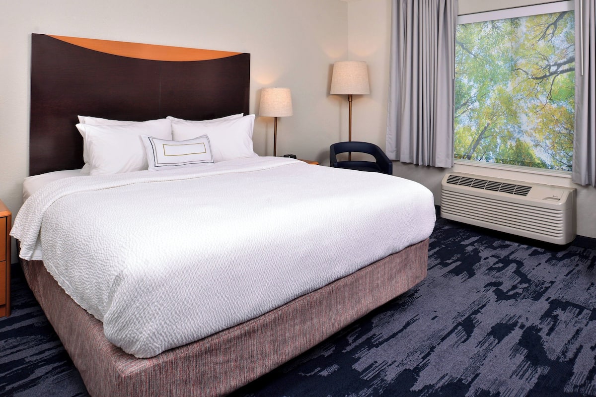 A hotel room with a large bed, two bedside lamps, a window with a view of trees, and a wall-mounted air conditioner.