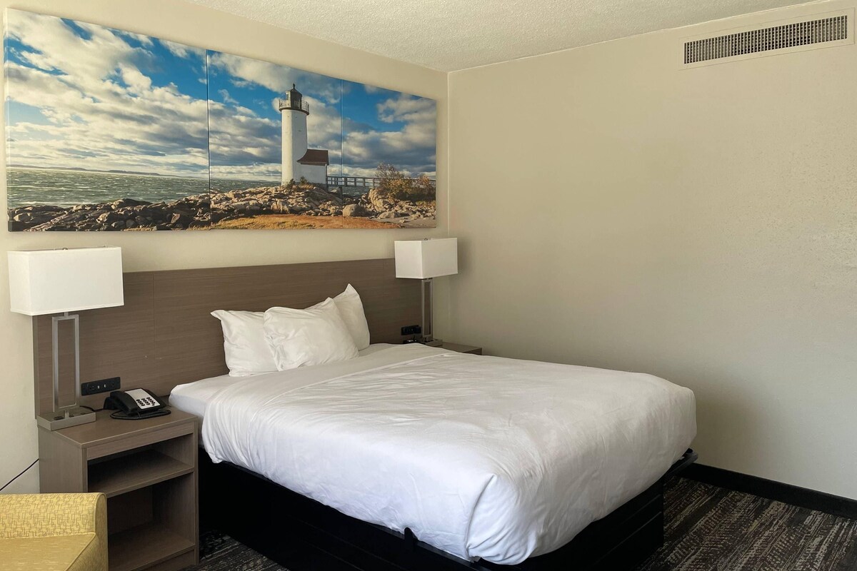 A hotel room with a bed, bedside lamps, a telephone, a small table, and a painting of a lighthouse.