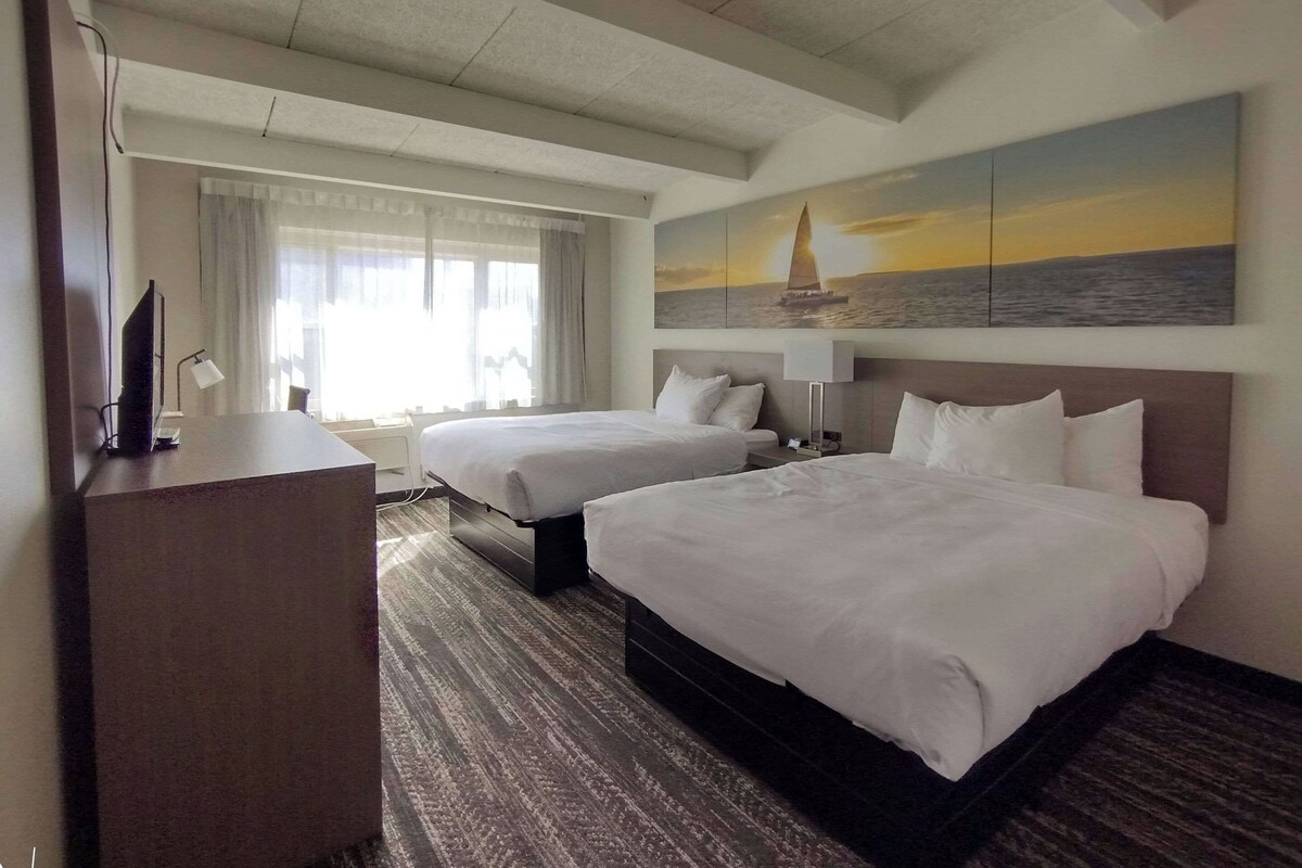 A hotel room with two beds, a desk, a chair, a television, and a large window with curtains.