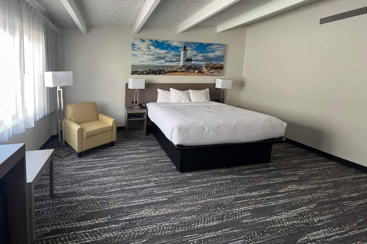 A hotel room with a bed, a chair, a desk, a lamp, and a painting on the wall.