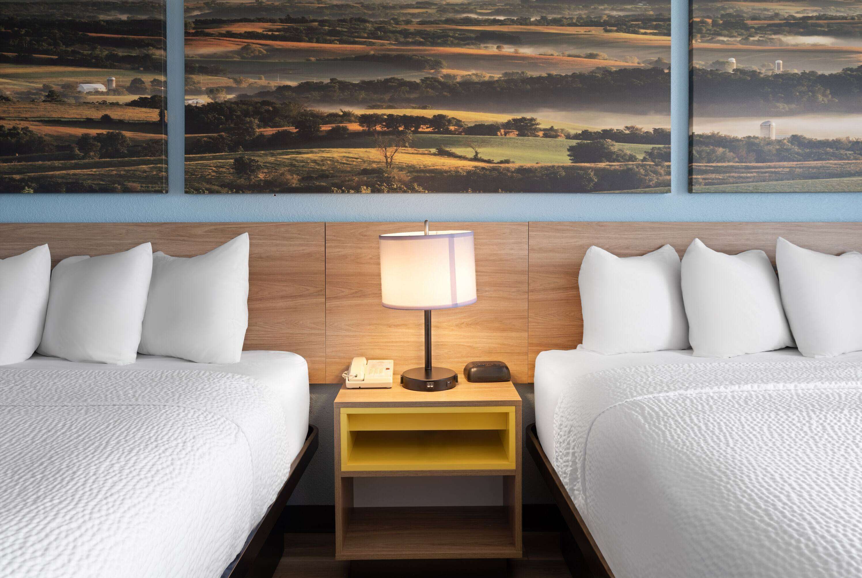A hotel room with two beds, a nightstand with a lamp, and a large landscape painting on the wall.