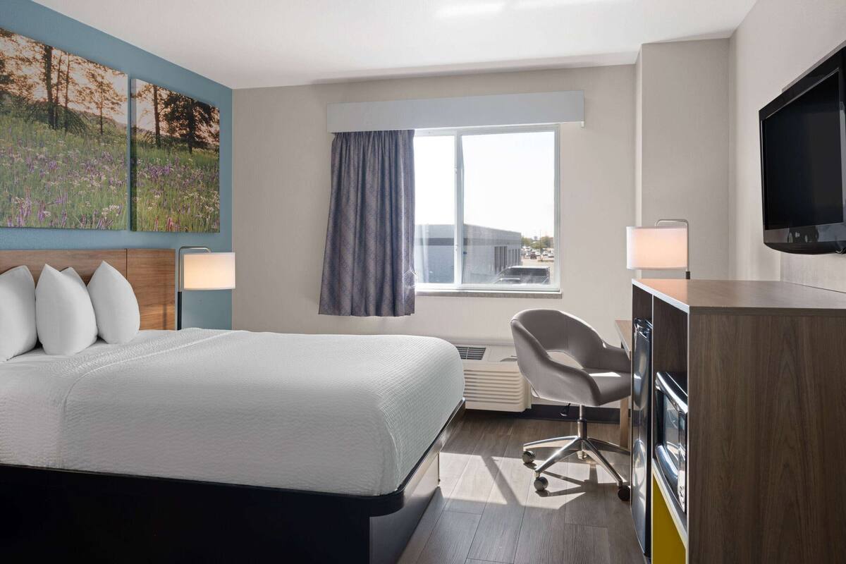 Habitación de hotel con cama, escritorio, silla, televisor y ventana con cortinas.