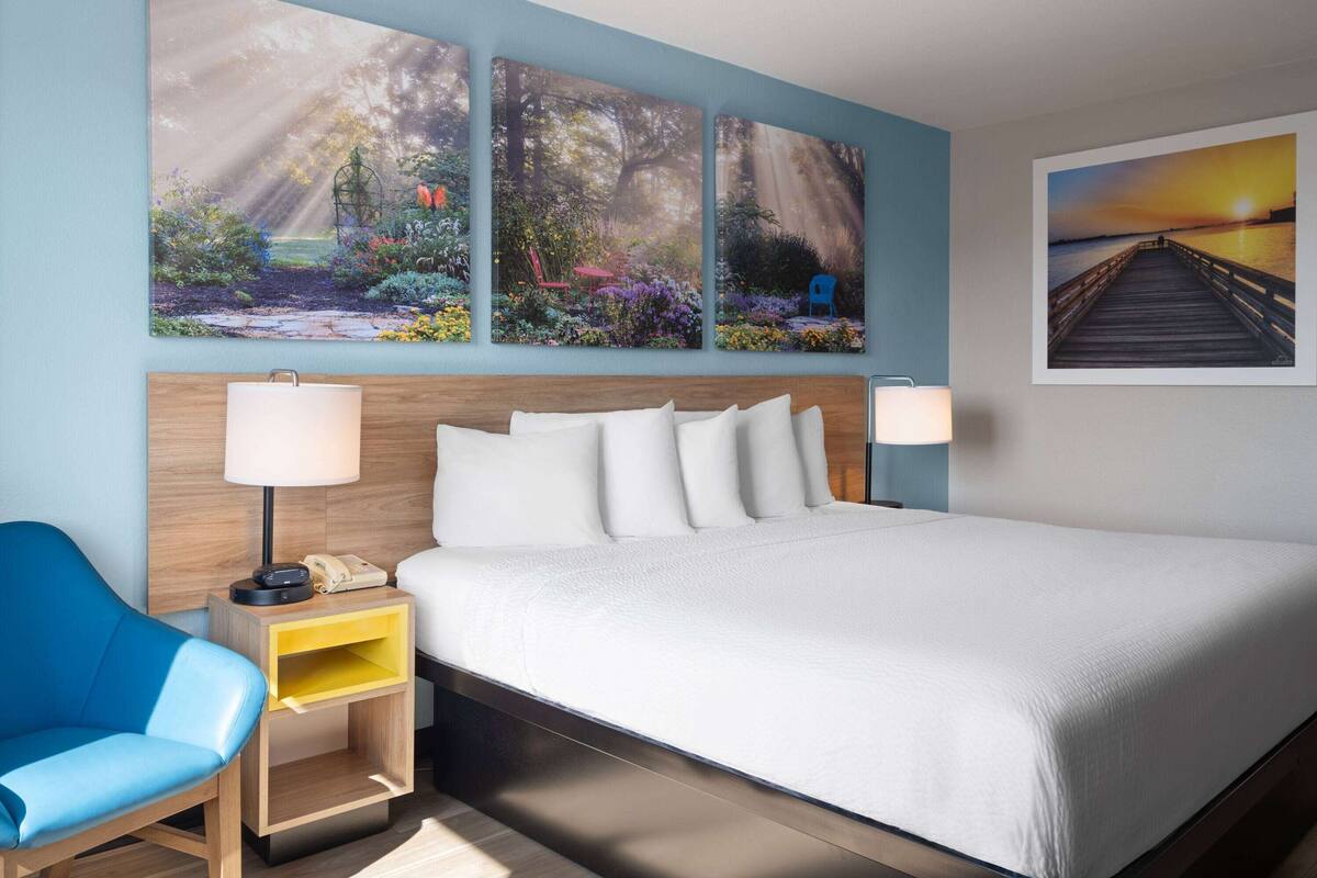 Una habitación de hotel con una cama grande, una silla azul, una mesita de noche de madera y tres cuadros enmarcados en la pared.