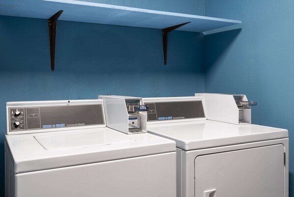 A laundry area with two white commercial washers and dryers, a blue wall, and a shelf above the machines.