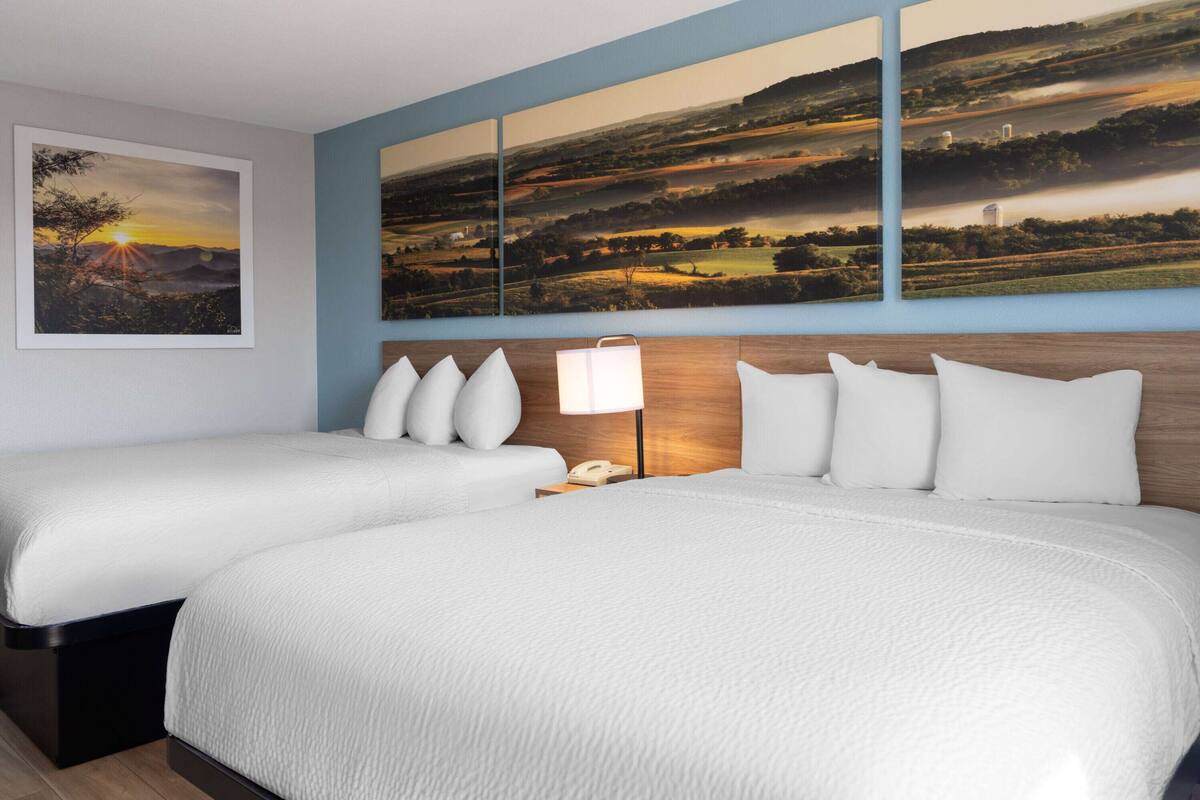 Habitación de hotel con dos camas, una mesita de noche con lámpara y un cuadro grande con un paisaje.