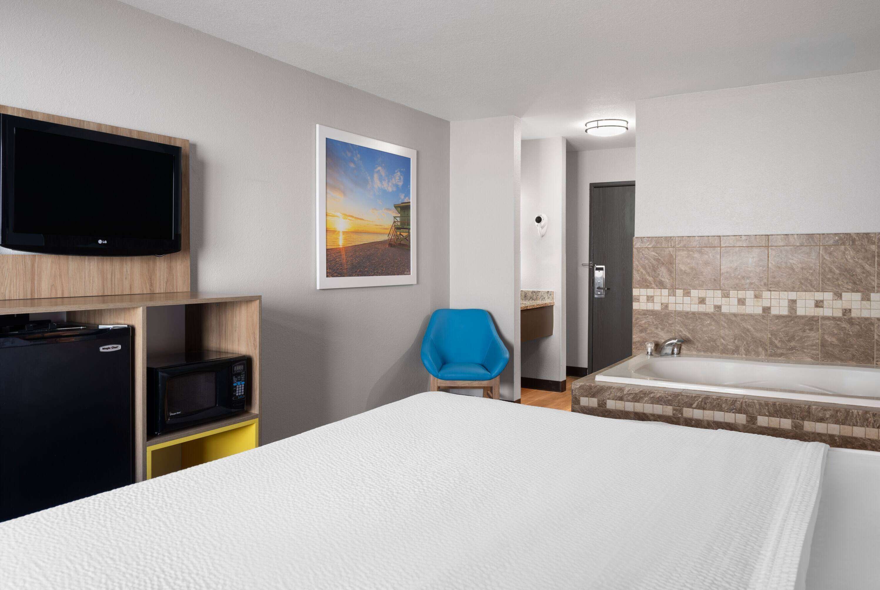 A hotel room with a bed, a TV mounted on the wall, a microwave, a blue chair, and a bathtub.