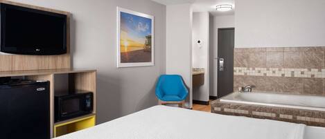 A hotel room with a bed, a TV mounted on the wall, a microwave, a blue chair, and a bathtub.