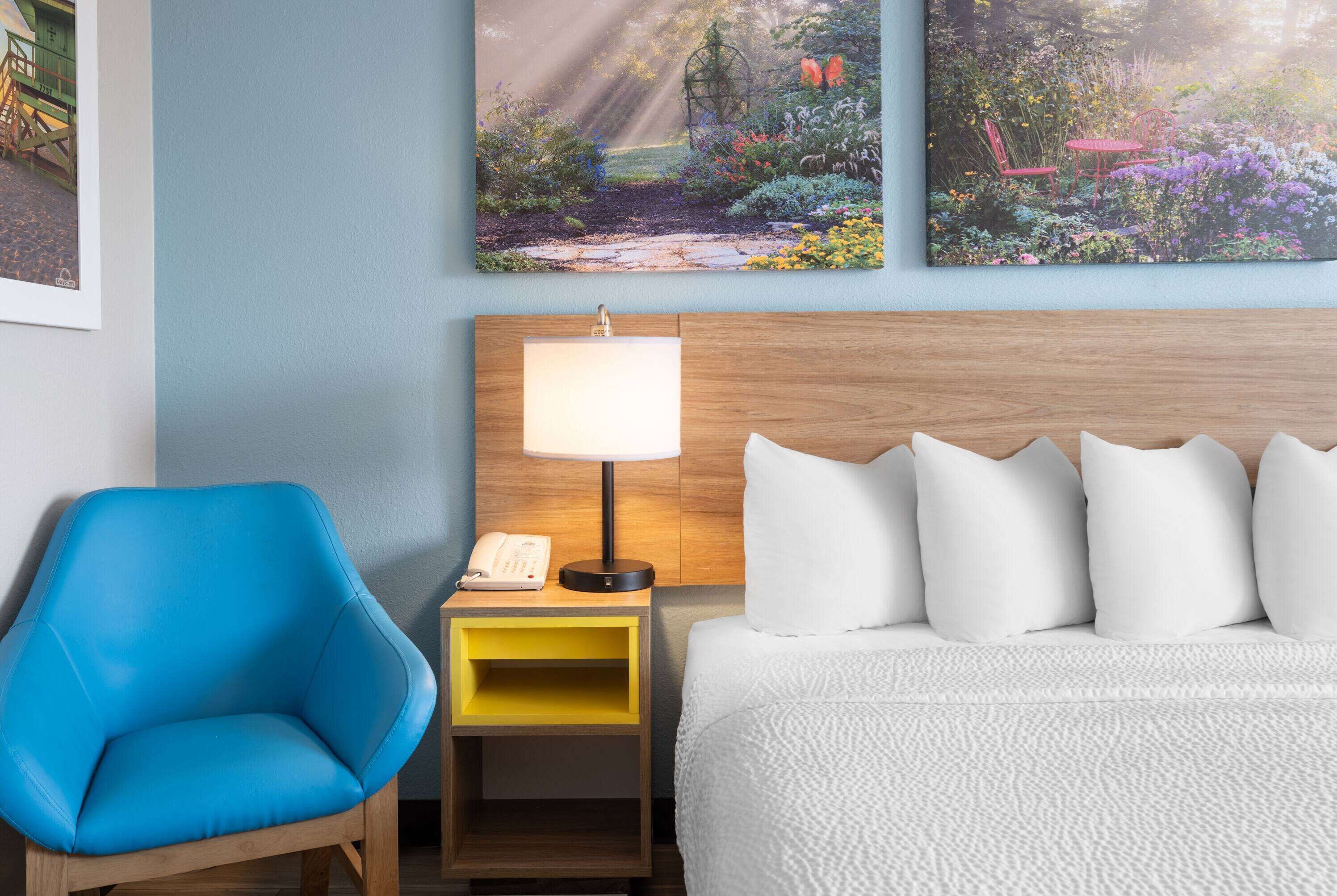 Ein Hotelzimmer mit einem Bett, einem Nachttisch mit Lampe, einem blauen Sessel und zwei Bildern an der Wand.